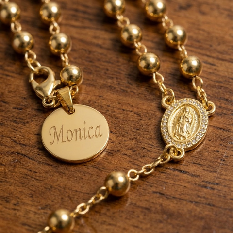 Close-up of gold bead rosary necklace with a round engraved name tag, clasp and Our Lady of Guadalupe medal with crystal border