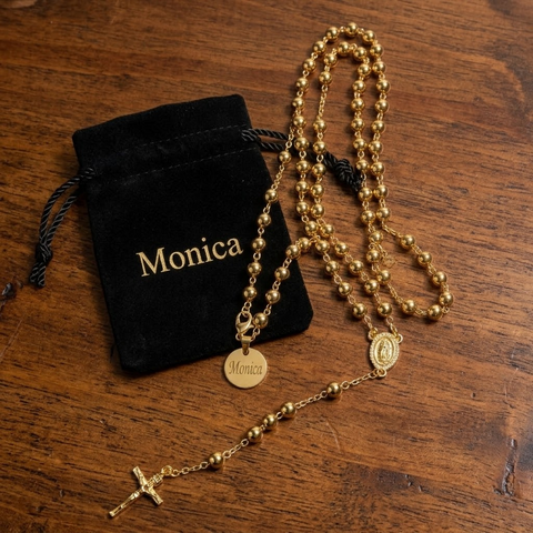 Personalized gold rosary necklace with an engraved Monica name tag, Our Lady of Guadalupe medal and crucifix beside a black velvet pouch