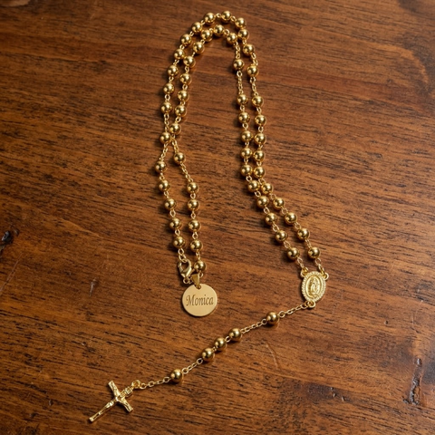 Gold bead rosary necklace with a round Monica name tag and crucifix laid flat on dark wood